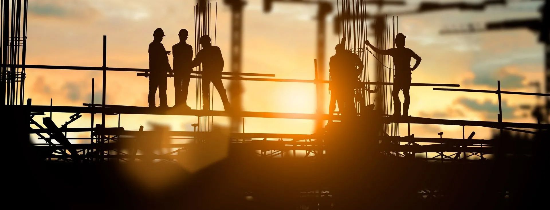 Silhouetten von Bauarbeitern mit Schutzhelmen auf einem Gerüst mit Bewehrungsstahl bei Sonnenuntergang auf einer Baustelle